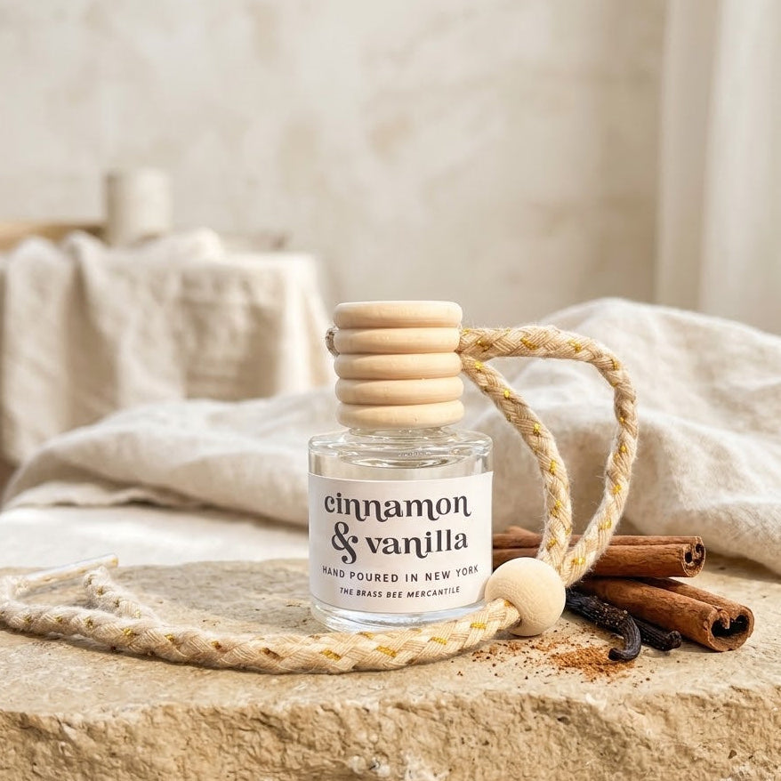 A small clear glass bottle labeled 'cinnamon & vanilla' with a light wood cap and braided rope handle, styled on a stone slab with cinnamon sticks and a bead in a softly lit indoor setting.